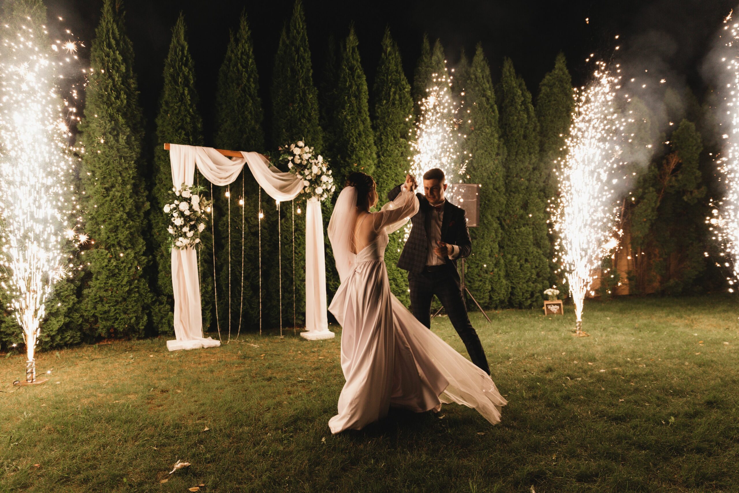 A couple enjoying a romantic dance at night surrounded by fireworks and fairy lights at their outdoor wedding.